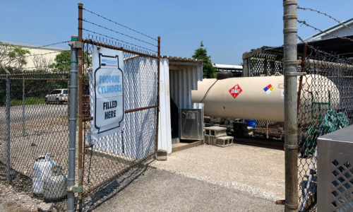 Outdoor propane exchange and refill station with signage indicating 'Propane Cylinders Filled Here' next to a fenced-in propane tank.